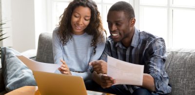 Married couple sitting on sofa at home paying bills online with online banking on laptop. Balancing bank account, feeling satisfied and happy.