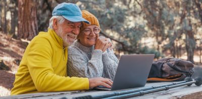 Head shot portrait close up of cute couple of old middle age people using computer pc outdoors sitting at a wooden table in the forest of mountain in nature with trees around them.