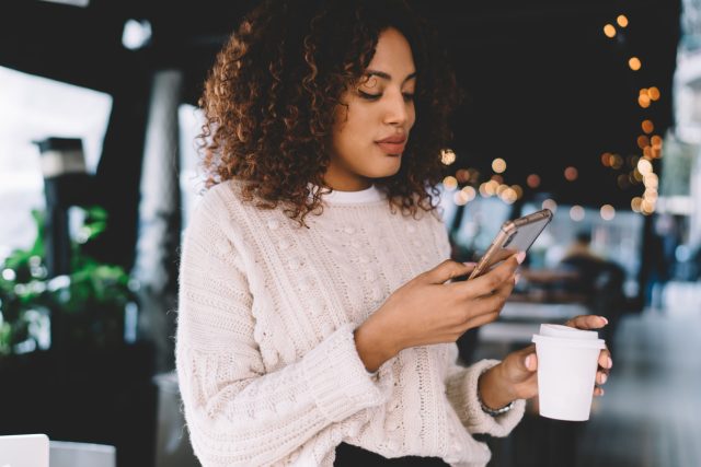 Smiling female holding a mobile phone using text banking to check balances while drinking cofffee.